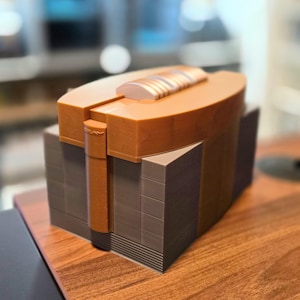 May include: A 3D-printed, geometric box in shades of brown and tan. The box has a layered design with a rounded top and a rectangular base. The top features a lighter-colored, curved accent. The box is sitting on a wooden surface.