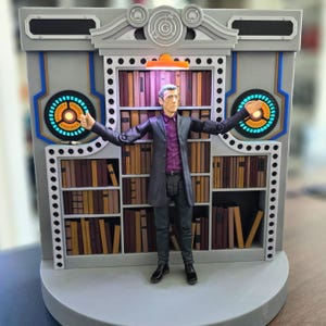 May include: A detailed action figure of a man in a long black coat and purple shirt, standing in front of a bookshelf. The bookshelf is part of a larger gray structure with blue and orange accents. The figure has its arms outstretched.