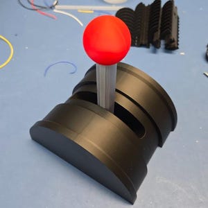 May include: A black joystick with a red ball-shaped top. The joystick has a cylindrical shaft and a base with a curved front. The image is taken from a slightly elevated angle, showing the details of the joystick's design.