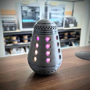 May include: A dark grey, cylindrical lamp with a textured surface and circular and hexagonal openings. The lamp has glowing pink lights visible through the openings. The lamp is on a wooden surface.