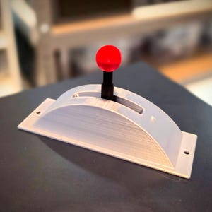 May include: A light grey, arched control lever housing with a central slot and a black vertical shaft topped with a bright red spherical knob. The base is rectangular with two small circular mounting holes on the flat ends for secure installation.