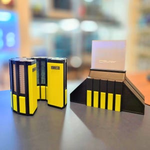 May include: A 3D-printed model of a Cray-2 supercomputer. The model features black and yellow components, with the text "CRAY-2" visible on the side. The model is on a gray surface.
