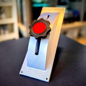 May include: A silver-colored metal mounting plate featuring a central vertical slot. A black, star-shaped adjustment knob with a bright red circular center is positioned within the slot, allowing for vertical movement along the plate's length.