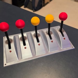 May include: A silver-colored control panel with five levers. Each lever has a black stem and a brightly colored spherical knob: two red, two yellow, and one red. The panel has small red lights near the base of each lever.