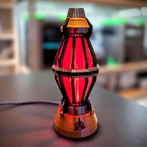 May include: A vertical, cylindrical lamp featuring a black frame with a glowing red interior. The base is tan with a black and red emblem. The structure has a metallic silver ring around the middle and a textured, tapered top section.