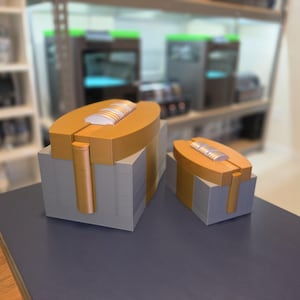 May include: Two 3D-printed objects. Each has a gold-coloured top with a white and silver accent, atop a stack of grey blocks. The larger piece has a gold-coloured cylindrical support. The objects are on a dark blue surface.