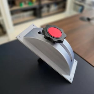 May include: A gray, angled bracket with a black and red knob. The bracket has a rectangular base with a curved top and two slots. The knob is black with a red center. The object is on a dark surface.
