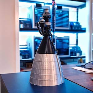 May include: A 3D printed model of a rocket engine with a black nozzle and a silver-colored body. The engine is sitting on a wooden table.