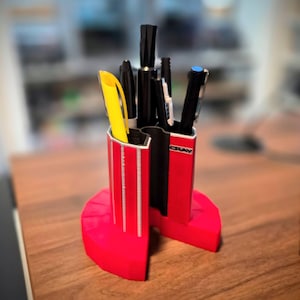May include: A red pen holder with a semi-circular base, holding various pens and markers. The pen holder has vertical silver stripes and the word "CRAY" on the side. A yellow pen is visible among the black pens.