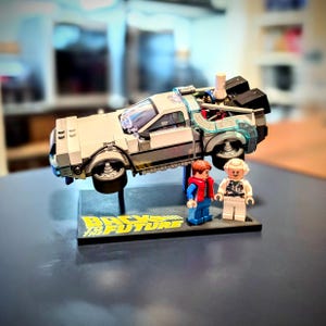 May include: A Lego DeLorean time machine model from the movie "Back to the Future" with two Lego figures. The car is silver with black accents and sits on a black base with the movie title in yellow and blue.
