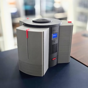 Könnte beinhalten: Ein grau-schwarzer Computer mit kreisförmiger Oberseite und kleinem blauem Bildschirm mit dem Wort "CRAY". Der Computer hat eine rechteckige Form mit roten Akzenten und vertikalen Linien an der Seite.