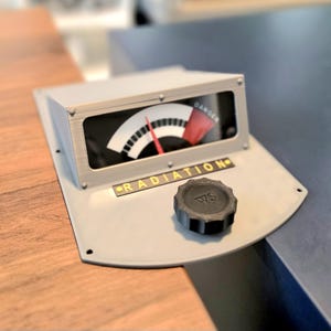 May include: A grey, retro-style radiation meter with a black and white gauge and a red needle pointing towards "Danger." The word "RADIATION" is printed in yellow. A black knob is on the base.