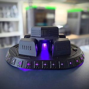 May include: A matte black, circular, disc-shaped object with a central raised structure featuring geometric panels and horizontal vents. The base is lined with a series of small, square, recessed ports that emit a bright, glowing purple light.
