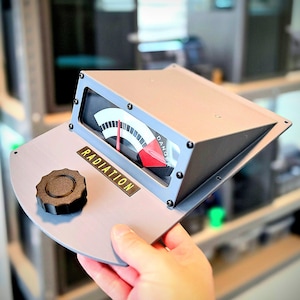 May include: A gray, wedge-shaped radiation gauge prop featuring a black circular knob, a rectangular display with a red needle pointing to a scale, and the word RADIATION printed in yellow text on a black background with the word DANGER visible.
