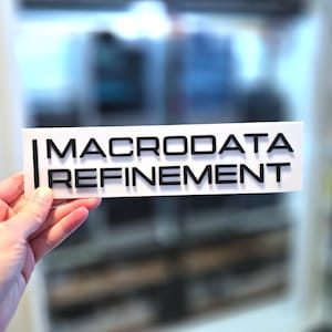 May include: A white rectangular sign with the words "MACRODATA REFINEMENT" in black, sans-serif font. The sign is held up in front of a blurred background.