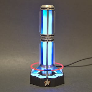 May include: A silver and black futuristic-looking lamp with a blue light and a red ring around the base. The lamp has a Starfleet logo on the base.