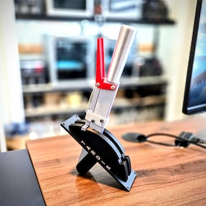 May include: A silver and black device with a red handle and the word "TARDIS" printed on the base. The device has a lever-like design with a spring and is mounted on a black base. The background is blurred, suggesting an office or workspace setting.
