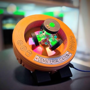 May include: A circular orange device with a central rotating mechanism featuring black discs, metal springs, and glowing green and pink lights. The frame displays engraved circular icons and a white rectangular label reading DEMATERIALIZATION.