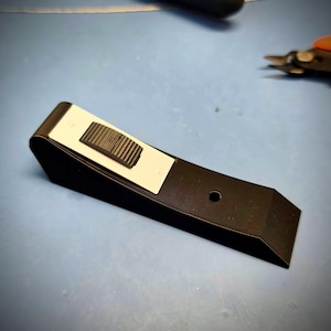 May include: A black wedge-shaped component featuring a rectangular white inset panel on its sloped top surface. A black textured thumbwheel is positioned within the white panel, and a small circular hole is located on the flat black base section.