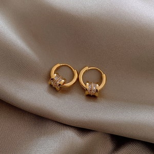 Könnte beinhalten: Ein Paar goldene Creolen mit einem klaren Kristall-Akzent. Die Ohrringe sind aus Metall und haben ein schlichtes, elegantes Design.