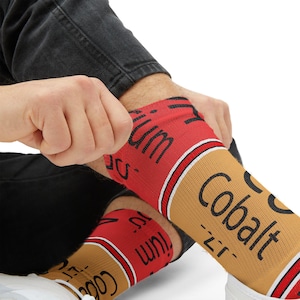 May include: A pair of knee-high socks with a red and tan design. The socks feature the word "Cobalt" and "Zirconium" printed on them. The socks are being pulled up over the ankles.