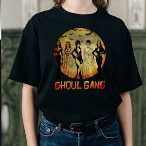 Może przedstawiać: Czarny T-shirt z grafiką przedstawiającą pięć kobiet przed pełnią księżyca z latającymi wok&oacute;ł nietoperzami. Pod księżycem nadrukowany jest napis "Ghoul Gang".