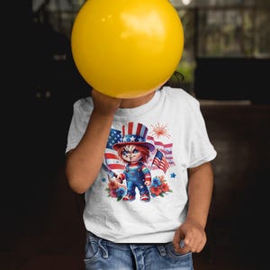 May include: White t-shirt featuring a graphic of a doll in a patriotic hat and overalls, holding a knife and an American flag. The design includes a backdrop of the American flag and fireworks. The shirt is worn by a person holding a yellow balloon.