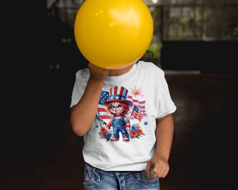 May include: White t-shirt featuring a graphic of a doll in a patriotic hat and overalls, holding a knife and an American flag. The design includes a backdrop of the American flag and fireworks. The shirt is worn by a person holding a yellow balloon.