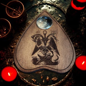 May include: A wooden planchette with a glass lens, featuring an engraved image of Baphomet. The planchette is surrounded by lit red candles and small wooden bowls. The planchette is a heart shape and is used for communicating with spirits.