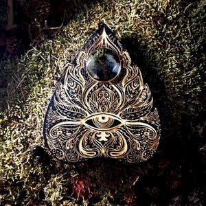 May include: A black and gold pendant with an eye symbol and detailed floral carvings. It has a large, dark, round stone at the top. The design is set against a mossy background.