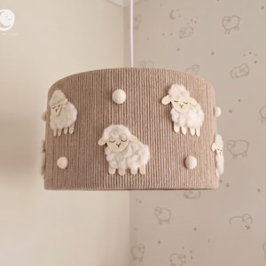 Könnte beinhalten: Ein zylindrischer beigefarbener Lampenschirm mit gerippter Textur, verziert mit dreidimensionalen, flauschigen weißen Schaf-Applikationen und kleinen weißen Pompons. Die Schafe haben einfache, liniengezeichnete Gesichter mit geschlossenen Augen.