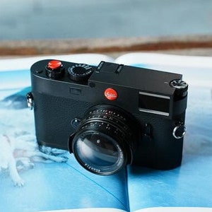 May include: A black Leica camera with a red dot on the top and a silver lens. The camera is sitting on a book with a blue and white image.