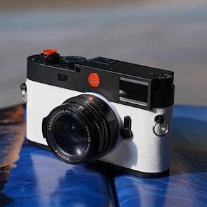 May include: A white and black Leica camera with a red dot logo on a blue and white book. The camera has a black lens and a silver ring around the lens.