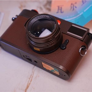 May include: A brown leather camera with a silver lens and a silver ring around the lens. The lens has the text "Leica Summicron-R 40mm f/2" on it. The camera has a silver strap loop on the side.