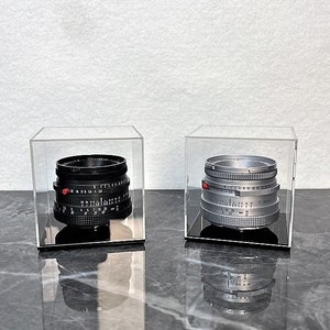 Puede incluir: Dos objetivos de cámara vintage en vitrinas de acrílico transparente. El objetivo de la izquierda es negro con un punto rojo y el objetivo de la derecha es plateado con un punto rojo.