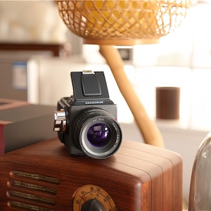 May include: A black and silver medium format camera with a large lens is positioned on top of a vintage wooden radio. The camera has a prominent lens and a viewfinder. The radio has a dark wood finish and a dial.