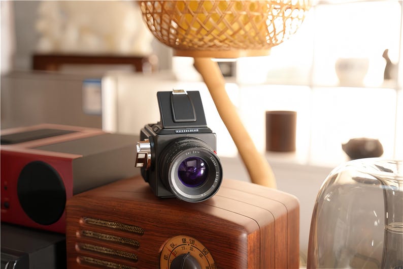 May include: A black and silver medium format camera with a large lens is positioned on top of a vintage wooden radio. The camera has a prominent lens and a viewfinder. The radio has a dark wood finish and a dial.