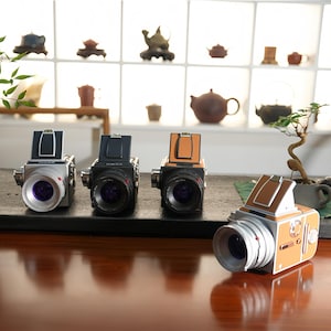 May include: Four vintage-style cameras with silver, black, and tan accents are arranged on a dark surface. The cameras have large lenses and a retro design. A decorative vase with greenery and a bonsai tree are also in the scene.