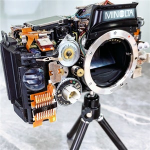 May include: A disassembled Minolta camera on a black tripod. The camera is missing its lens and has exposed internal components, including gears, wires, and a circuit board. The camera is labeled "MINOLTA" on the top.