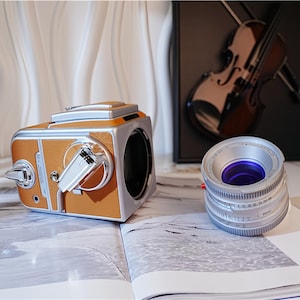May include: A vintage-style camera with a brown leather-like exterior and silver accents sits on an open book. A silver camera lens with a purple interior is next to the camera. The scene includes a violin and a speaker.