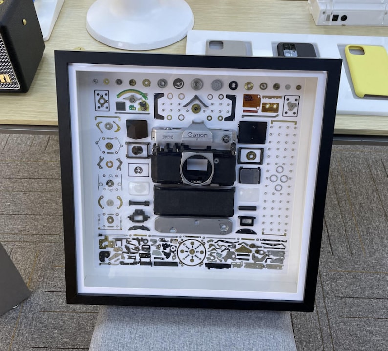 Puede incluir: Una imagen enmarcada en negro con fondo blanco que muestra una c&aacute;mara Canon desmontada con varias piezas y partes. La c&aacute;mara es plateada y negra con la palabra "Canon" impresa en ella.
