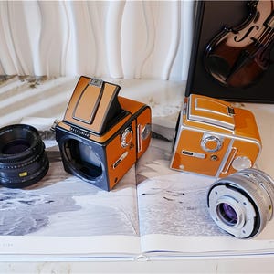 May include: Two orange and black vintage cameras with lenses are displayed on an open book. The cameras have a retro design with a tan top and silver accents. A black lens and a silver lens are also visible.