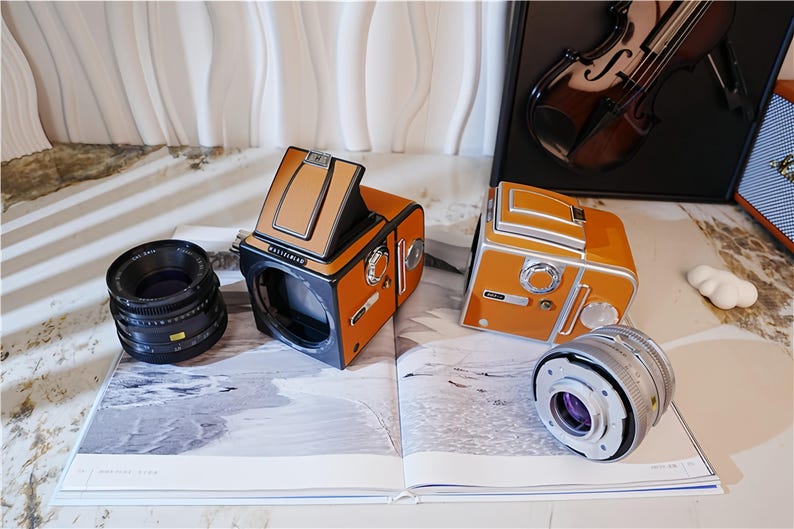 May include: Two orange and black vintage cameras with lenses are displayed on an open book. The cameras have a retro design with a tan top and silver accents. A black lens and a silver lens are also visible.