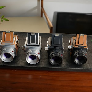 May include: Four vintage-style cameras with silver bodies and leather accents are displayed. Two have brown leather, one has black, and one has silver. Each camera has a prominent lens and a top-mounted viewfinder.