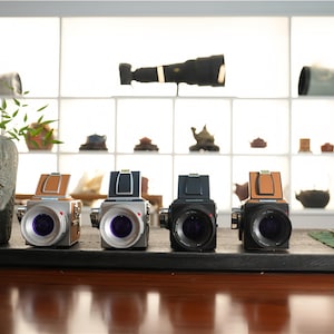 May include: Four vintage-style cameras with silver and black bodies, and brown or black top flaps, are displayed on a dark surface. The cameras have purple lenses. A decorative stone vase and bonsai tree are also present.