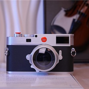 Op de afbeelding: Een zilveren en zwarte meetzoekercamera met een behuizing van gestructureerd kunstleer, een ronde lens met zilveren behuizing, een rood rond logo aan de voorzijde, een zilveren bovenplaat met een rode ontspanknop en metalen riemogen aan beide zijden.