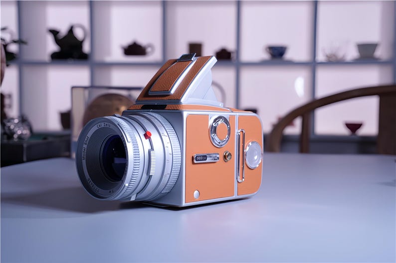 May include: A vintage-style medium format camera in orange and silver. The camera features a large lens, a leather top, and a retro design. The camera is sitting on a white surface.