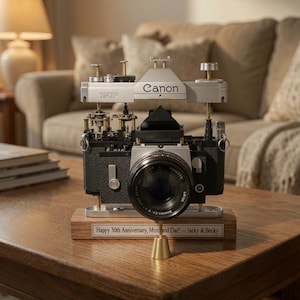 May include: A disassembled Canon FP camera mounted on a wooden base. The top housing is elevated, revealing internal gears and mechanisms. A lens is attached to the front. A metal plaque on the base reads: Happy 30th Anniversary, Mom and Dad! – Jacky & Becky.