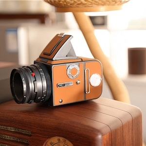May include: A vintage-style camera with a brown and orange body, black lens, and silver accents. The camera is placed on top of a wooden radio. The camera has a viewfinder and a lens with a red indicator.