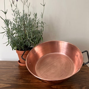 May include: A large copper pan with two black handles. The pan is sitting on a wooden table.